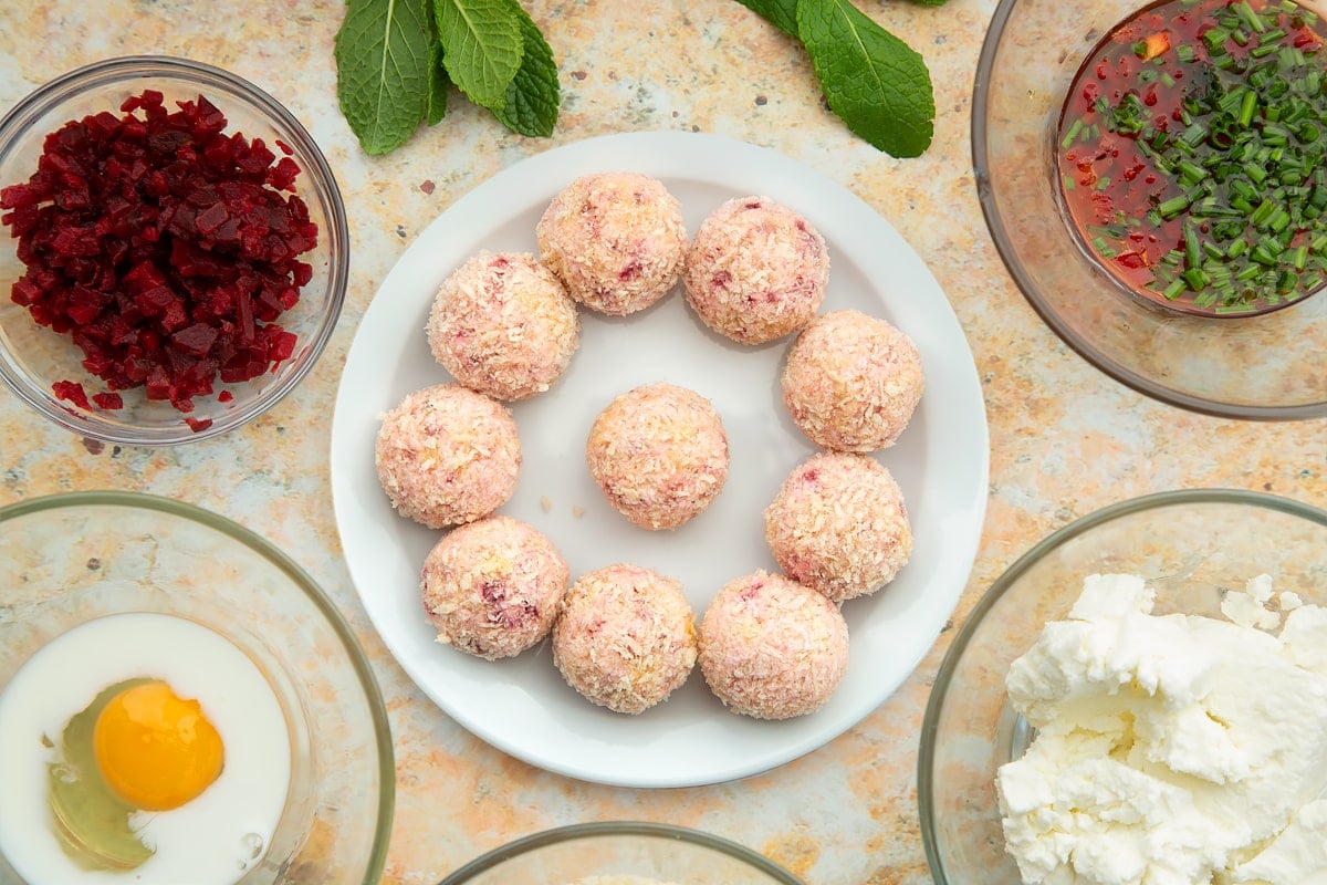 Goat's cheese and beetroot balls coated in breadcrumbs on a small white plate. Ingredients to make goat's cheese croquettes surround the plate.