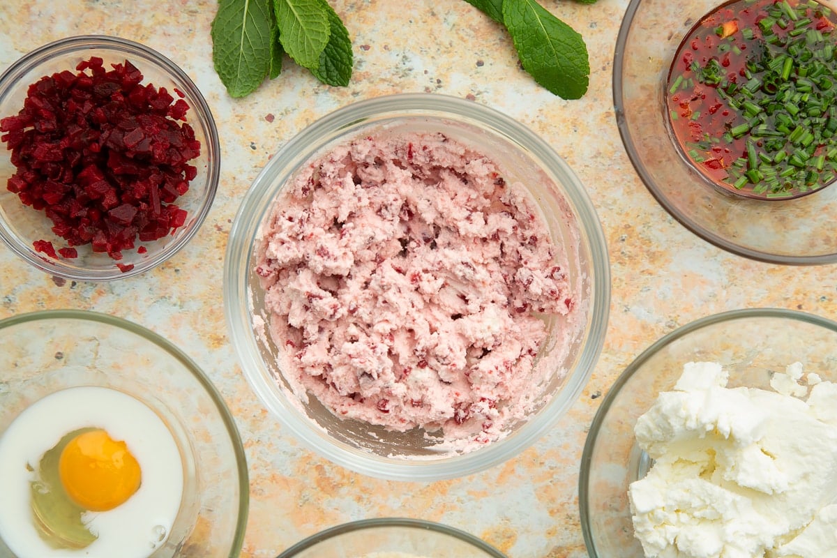 Mashed goat's cheese mixed together with chopped beetroot in a mixing bowl. Ingredients to make goat's cheese croquettes surround the bowl.