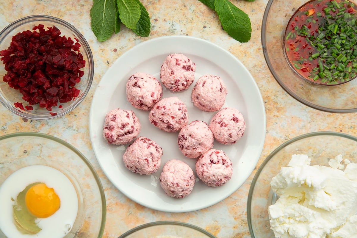 Goat's cheese and beetroot balls on a small white plate. Ingredients to make goat's cheese croquettes surround the plate.