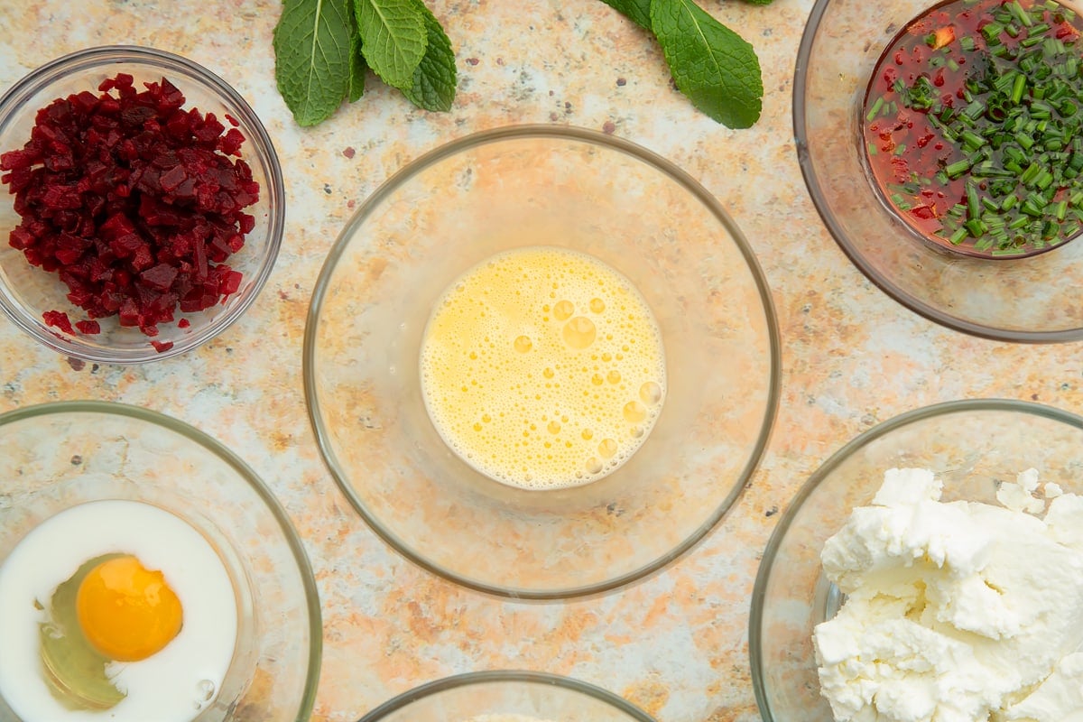 Egg and milk whisked together in a small glass bowl. Ingredients to make goat's cheese croquettes surround the bowl.