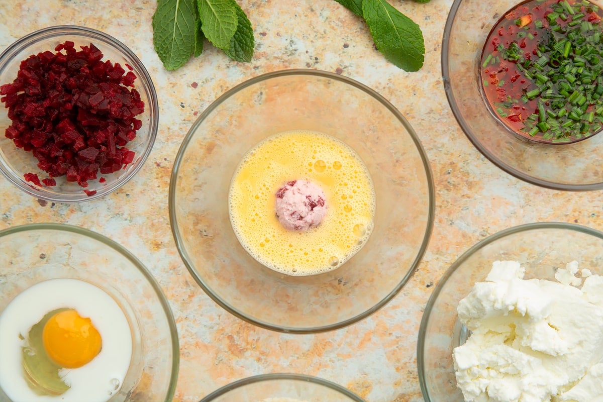 Egg and milk mix in a small glass bowl with a ball of goat's cheese and beetroot in the centre. Ingredients to make goat's cheese croquettes surround the bowl.