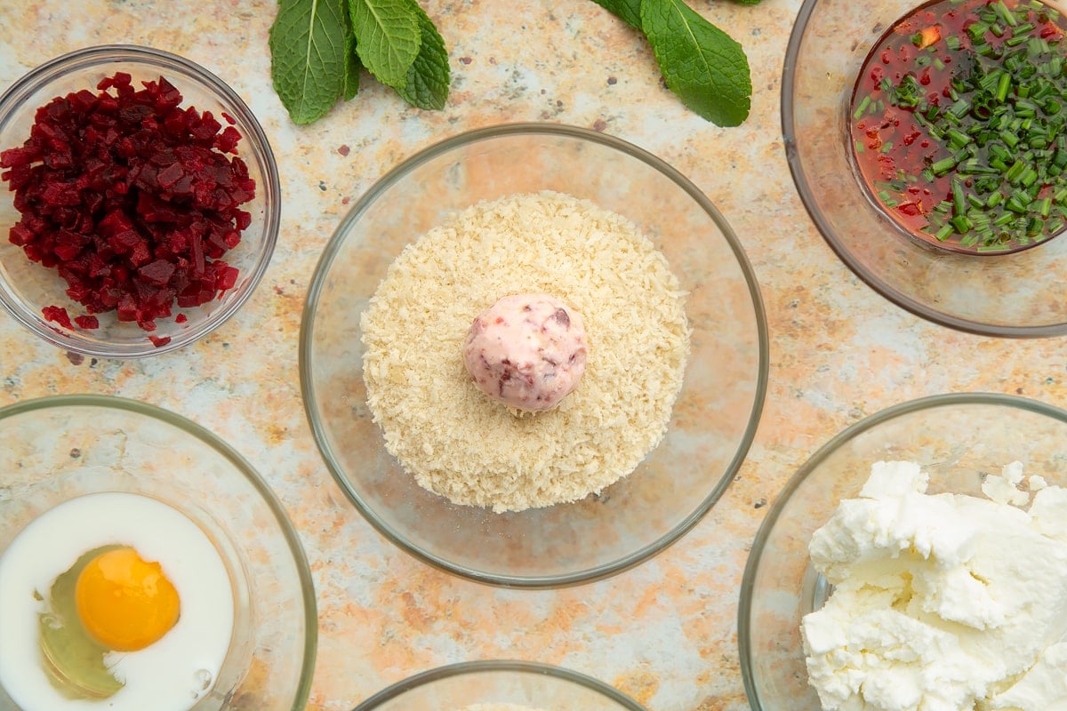 Breadcrumbs in a small glass bowl with a ball of goat's cheese and beetroot in the centre. Ingredients to make goat's cheese croquettes surround the bowl.