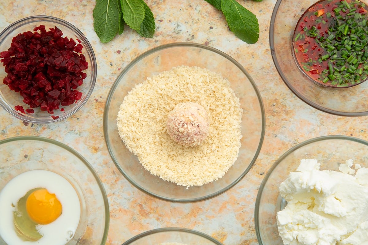 Breadcrumbs in a small glass bowl with a goat's cheese croquette in the centre. Ingredients to make goat's cheese croquettes surround the bowl.