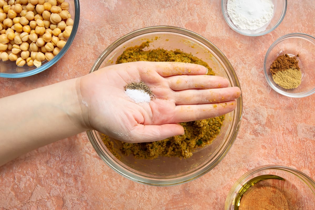 A hand holding a pinch of salt and pepper over a bowl of harissa falafel paste.