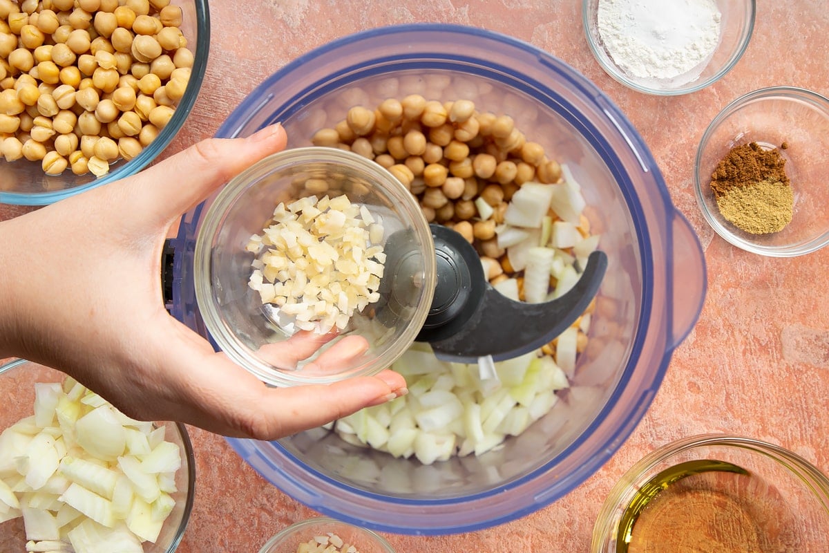 A hand holds a small bowl of chopped garlic over a food processor containing chickpeas and chopped onion for harissa falafel.