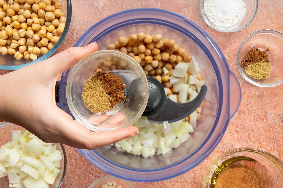 A hand holds a small bowl of spices over a food processor containing chickpeas, garlic and chopped onion for harissa falafel.