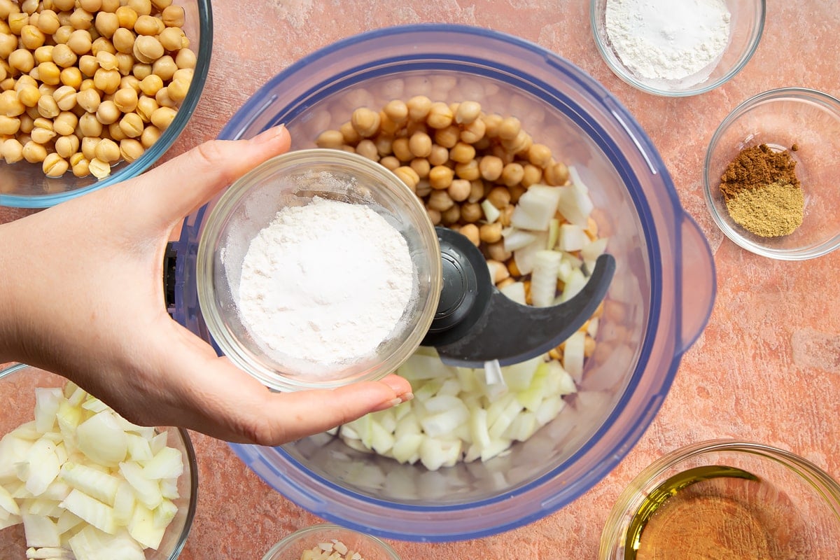 A hand holds a small bowl of flour and baking powder over a food processor containing chickpeas, spices, garlic and chopped onion for harissa falafel.