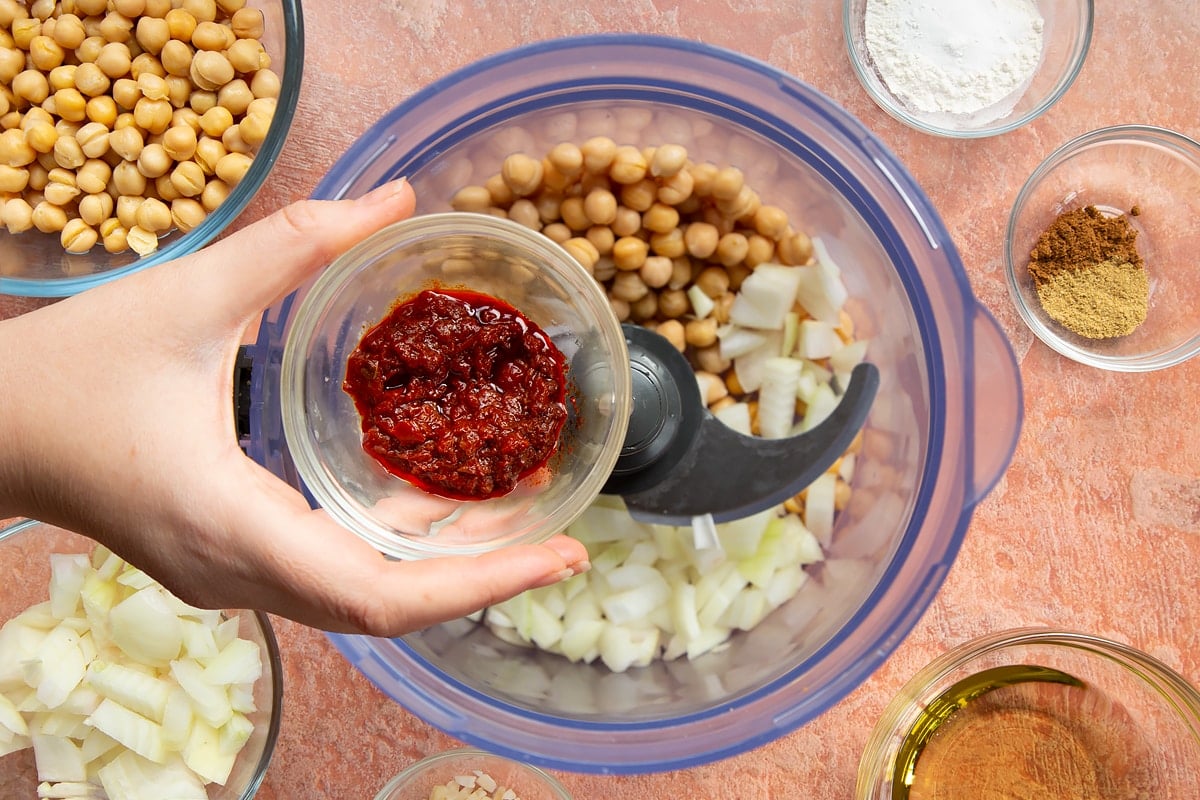 A hand holds a small bowl of harissa paste over a food processor containing chickpeas, spices, garlic, flour, baking powder and chopped onion for harissa falafel.