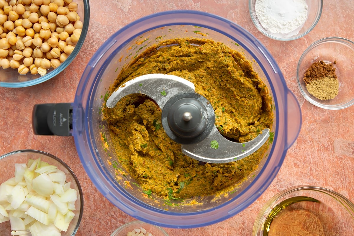 A food processor containing a harissa falafel paste made from chickpeas, spices, garlic, flour, baking powder, chopped onion, fresh parsley and coriander.