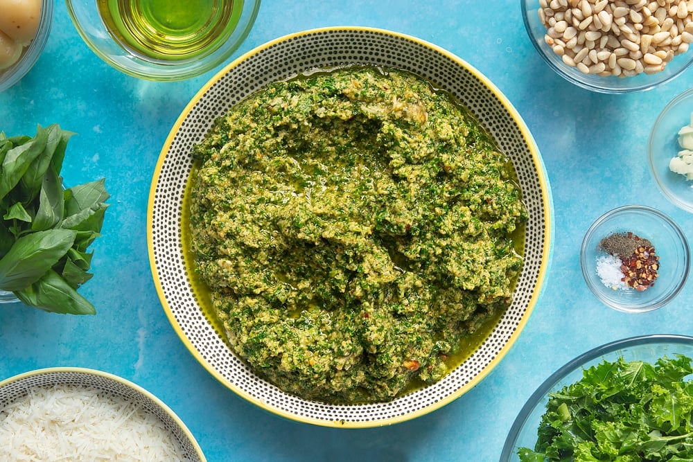 A bowl containing freshly made kale pesto. The bowl is surrounded by ingredients for kale pesto linguine.