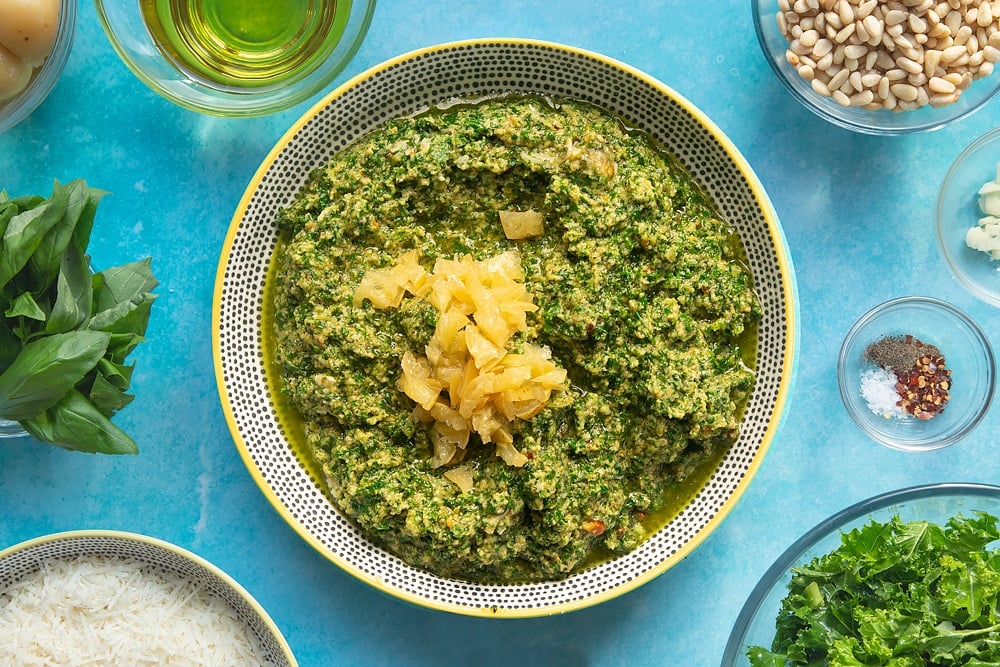 A bowl containing freshly made kale pesto with chopped preserved lemons on top. The bowl is surrounded by ingredients for kale pesto linguine.