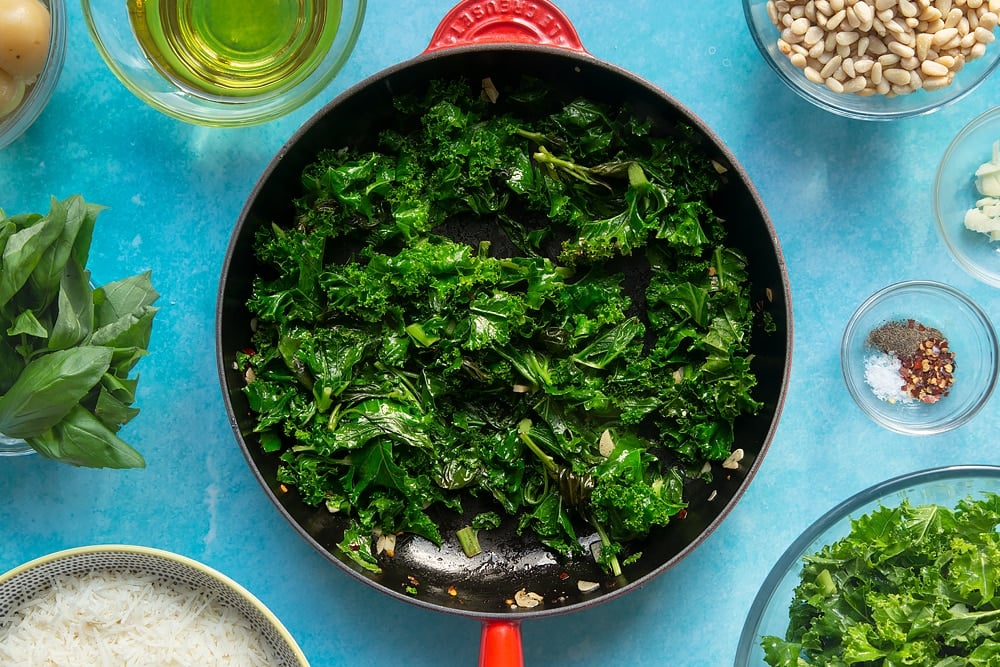 A pan containing wilted kale and basil, fried garlic, chilli salt and pepper. The pan is surrounded by ingredients for kale pesto linguine.