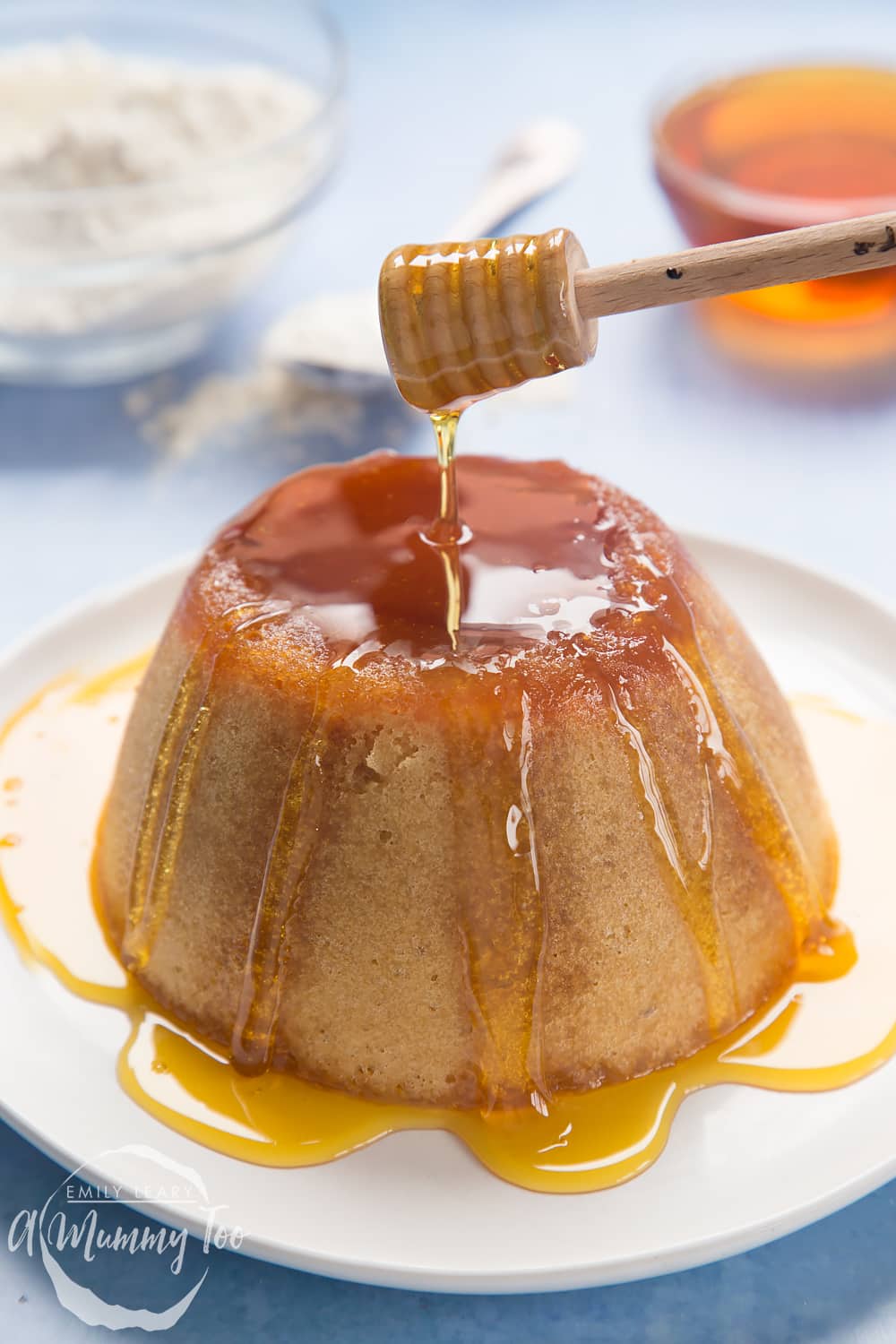 Front angle shot of honey dipper above Microwave syrup sponge pudding served in a white plate  with a mummy too logo in the lower-left corner