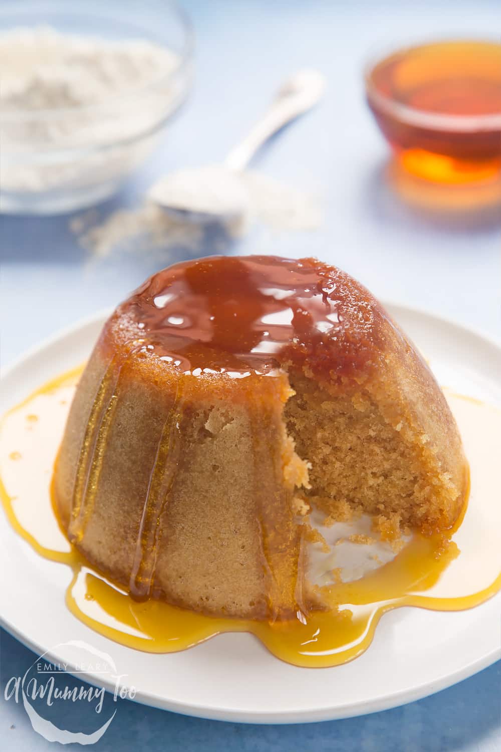 Front angle shot of syrup sponge pudding served in a white plate with honey with a mummy too logo in the lower-left corner