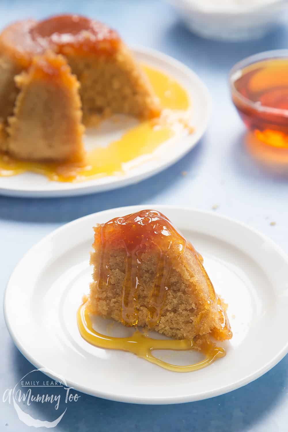 Front angle shot of a slice of sponge pudding with honey served in a white plate with honey with a mummy too logo in the lower-left corner