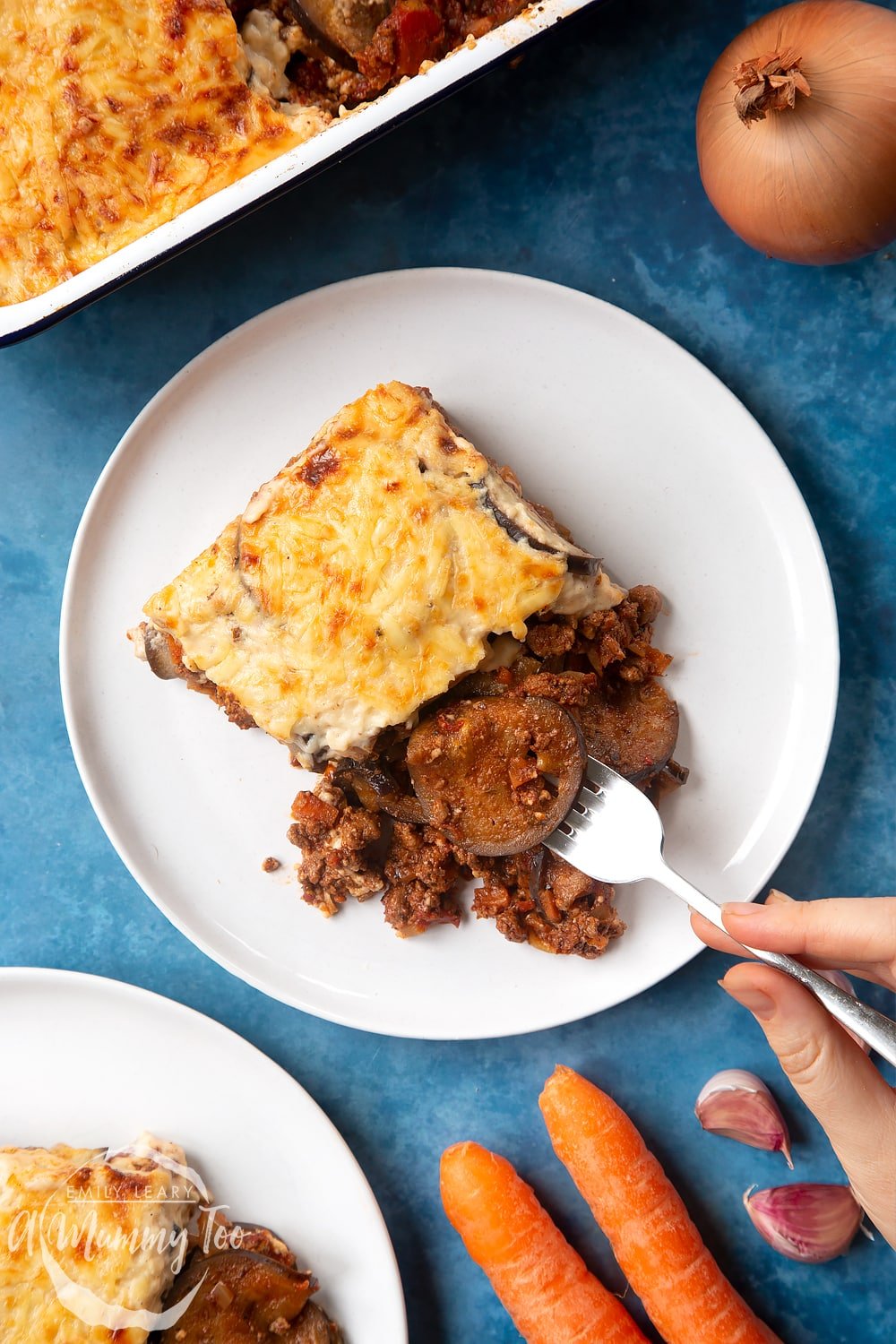 Overhead shot of a hand holding a fork picking up aubergine from Quorn Meat Free Mince Moussaka served on a white plate with logo in the lower-left corner