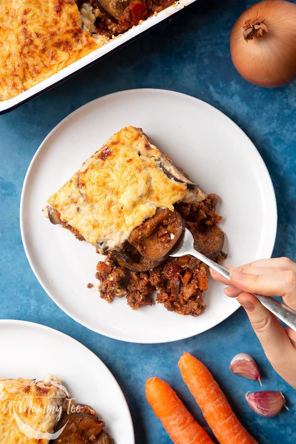 Overhead shot of a hand holding a fork picking up an aubergine from Quorn Meat Free Mince Moussaka served on a white plate