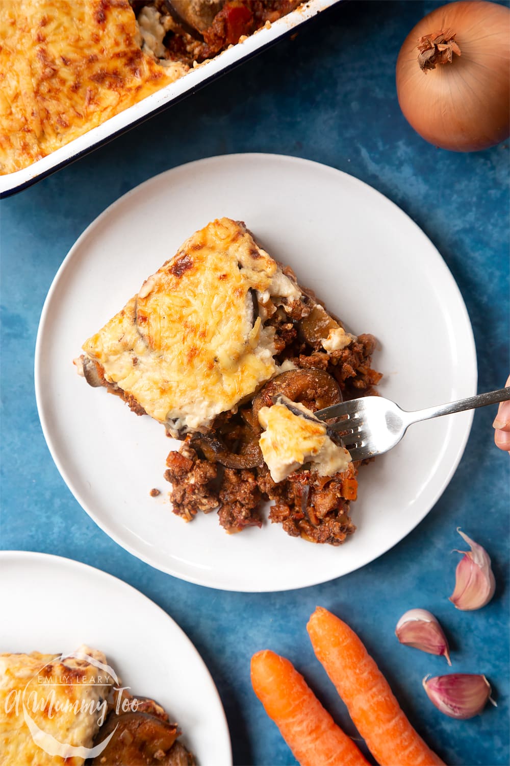 Overhead shot of a hand holding a fork picking up an aubergine from Quorn Meat Free Mince Moussaka served on a white plate