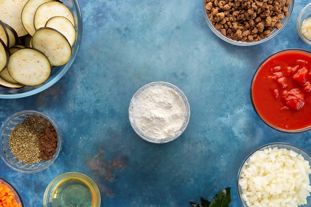 Overhead shot of cornflour in a small clear bowl surrounded by aubergine minced meat spices onion oil carrot garlic bayleaves and chopped tomatoes