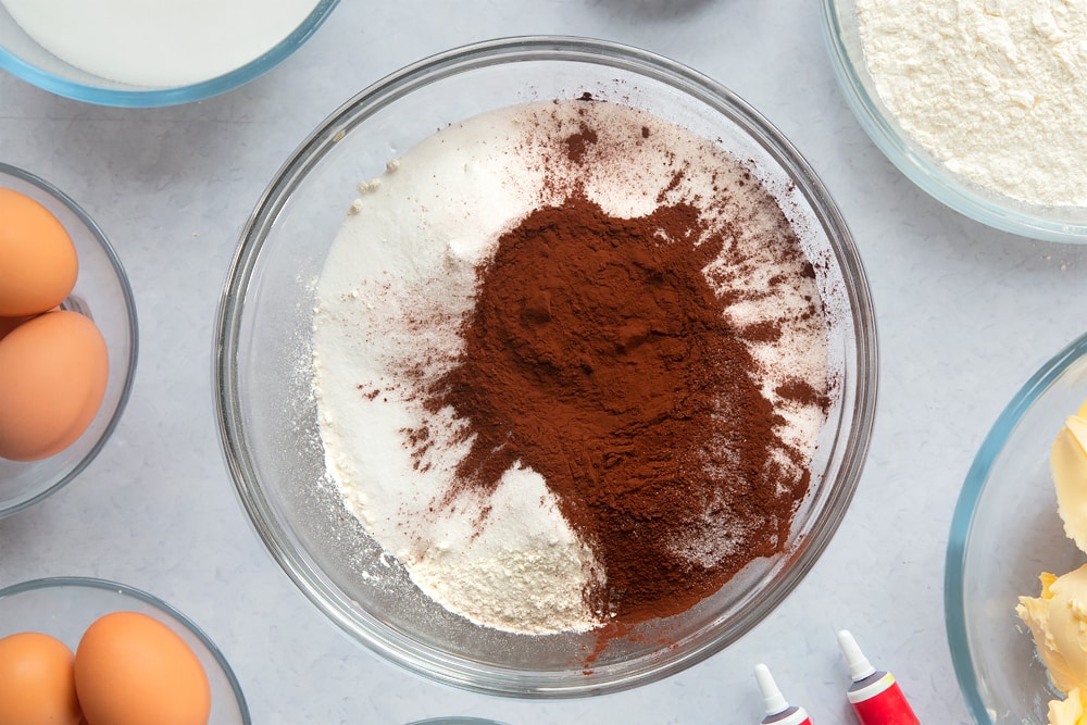 Flour, sugar and cocoa in a large glass mixing bowl. Ingredients to make filled red velvet cake surround the bowl. 