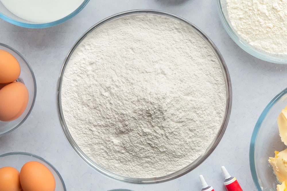 Cream cheese frosting with more icing sugar added in a large glass mixing bowl. Ingredients to make filled red velvet cake surround the bowl. 