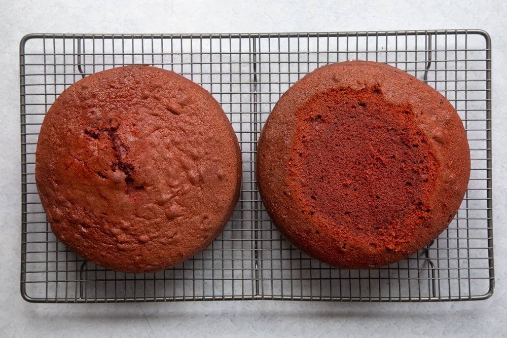 Two cooked red velvet sponges on. a cooling rack. One has had its top trimmed to create a flat surface.