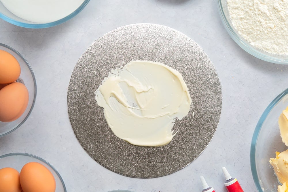 A silver cake board with a little cream cheese frosting spread on it. Ingredients to make filled red velvet cake surround the board. 