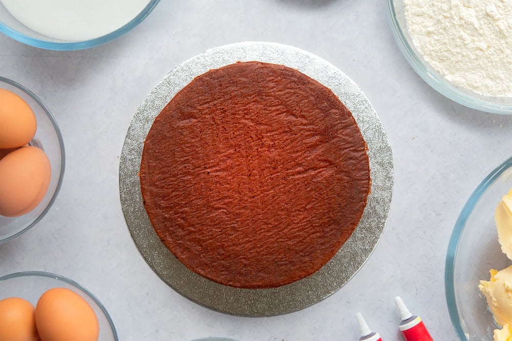 A silver cake board with a red velvet sponge on it. Ingredients to make filled red velvet cake surround the board. 