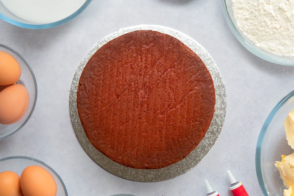 A silver cake board with a red velvet cake on it, sandwiched with cream cheese frosting. Ingredients to make filled red velvet cake surround the board. 