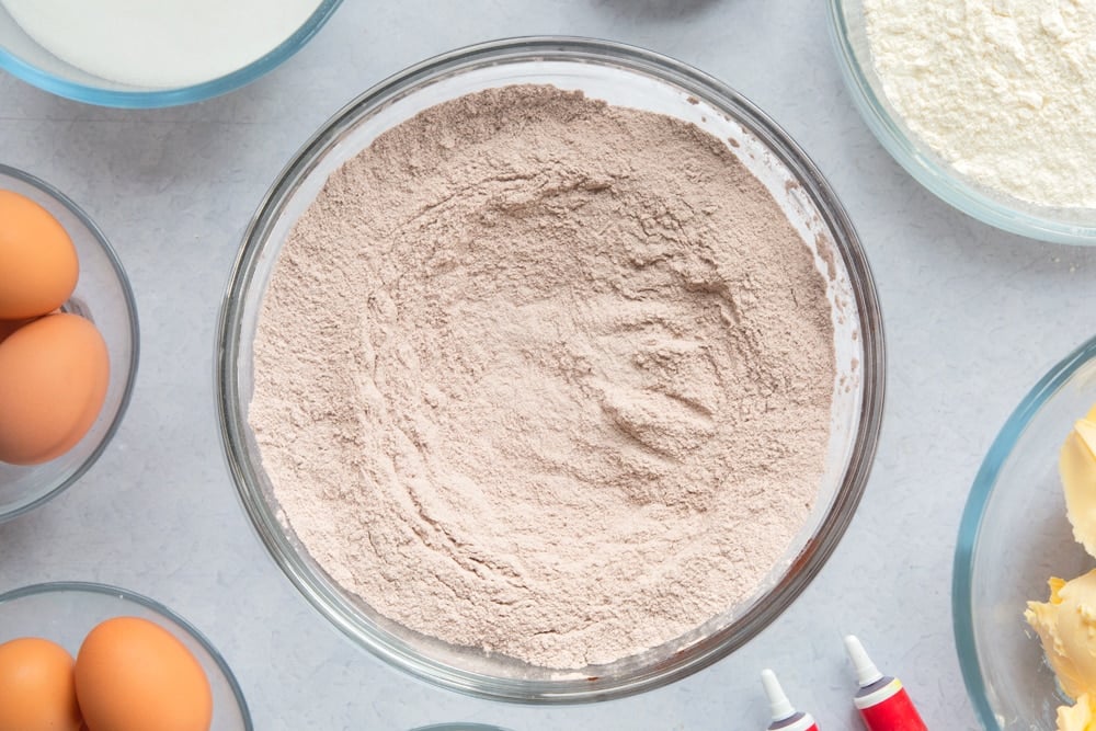 Flour, sugar and cocoa whisked together in a large glass mixing bowl. Ingredients to make filled red velvet cake surround the bowl. 