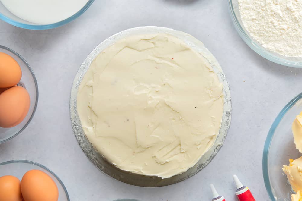 A silver cake board with a red velvet cake on it, covered with cream cheese frosting. Ingredients to make filled red velvet cake surround the board. 