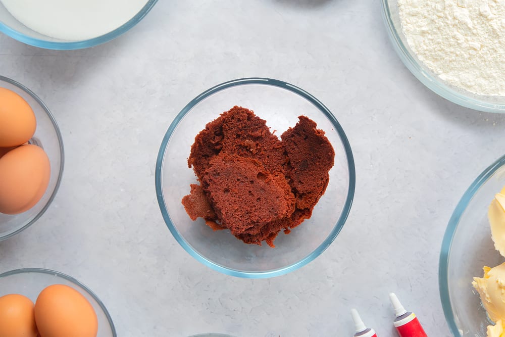 Red velvet cake offcuts in a small bowl. Ingredients to make filled red velvet cake surround the bowl. 