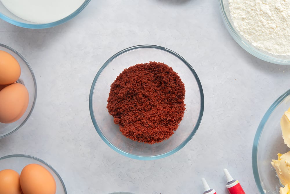 Red velvet cake crumbs in a small bowl. Ingredients to make filled red velvet cake surround the bowl. 