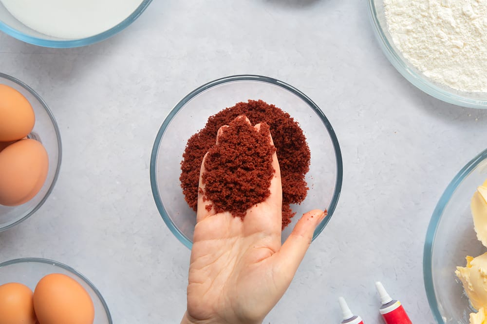 Red velvet cake crumbs in a small bowl. A hand holds some of the crumbs. Ingredients to make filled red velvet cake surround the bowl. 