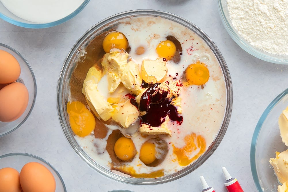 Flour, sugar, cocoa, margarine, eggs, milk and red food colouring in a large glass mixing bowl. Ingredients to make filled red velvet cake surround the bowl. 