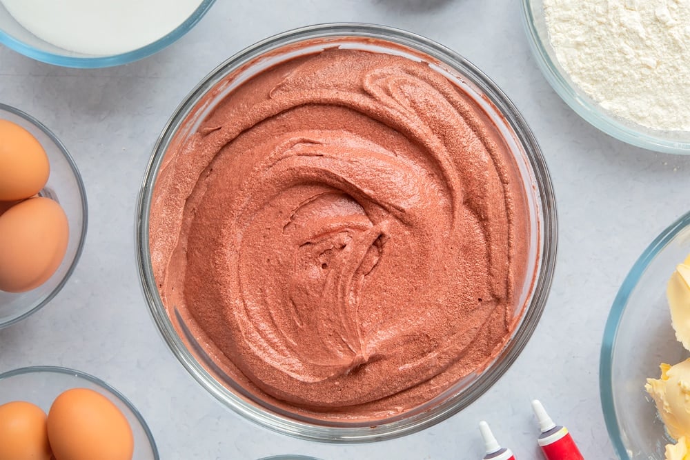 Red velvet cake batter in a large glass mixing bowl. Ingredients to make filled red velvet cake surround the bowl. 