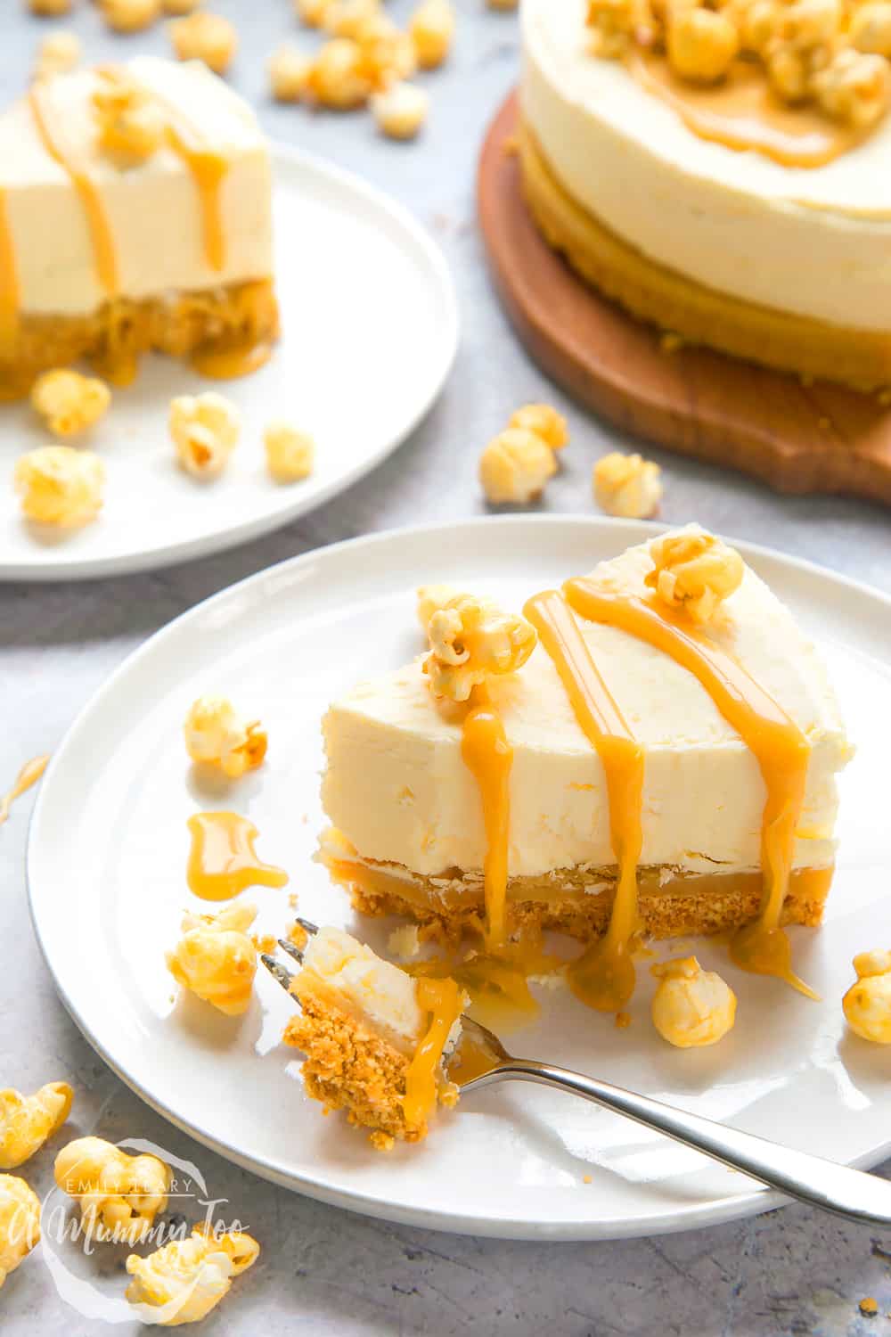 a slice of no-bake salted caramel cheesecake drizzled with caramel sauce and topped with popcorn on a white plate and a fork cutting a piece away.