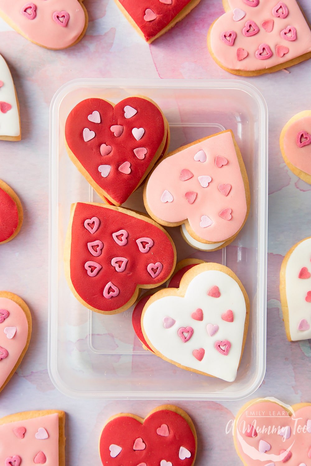 Overhead shot of valentines day cookies stacked in a clear plastic container with A Mummy Too logo in the bottom right hand corner