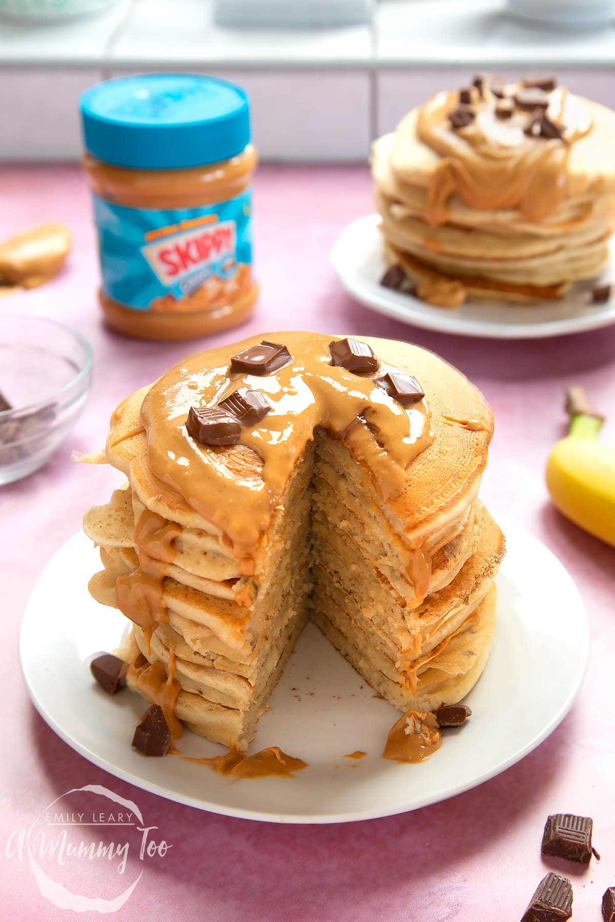 A stack of vegan peanut butter pancakes on a white plate. The stack is drizzled with more peanut butter and scattered with chunks of vegan chocolate. A large slice has been taken out of the whole stack, revealing the fluffy inner.