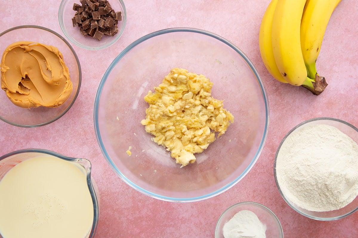 A bowl of mashed banana, surrounded by the ingredients for vegan peanut butter pancakes.