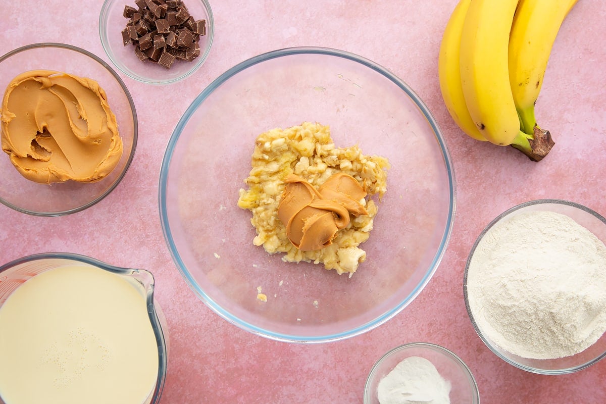 A bowl of mashed banana topped with smooth peanut butter, surrounded by the ingredients for vegan peanut butter pancakes.