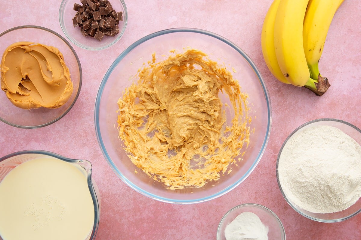 A bowl of banana and smooth peanut butter whisked together. The bowl is surrounded by the ingredients for vegan peanut butter pancakes.