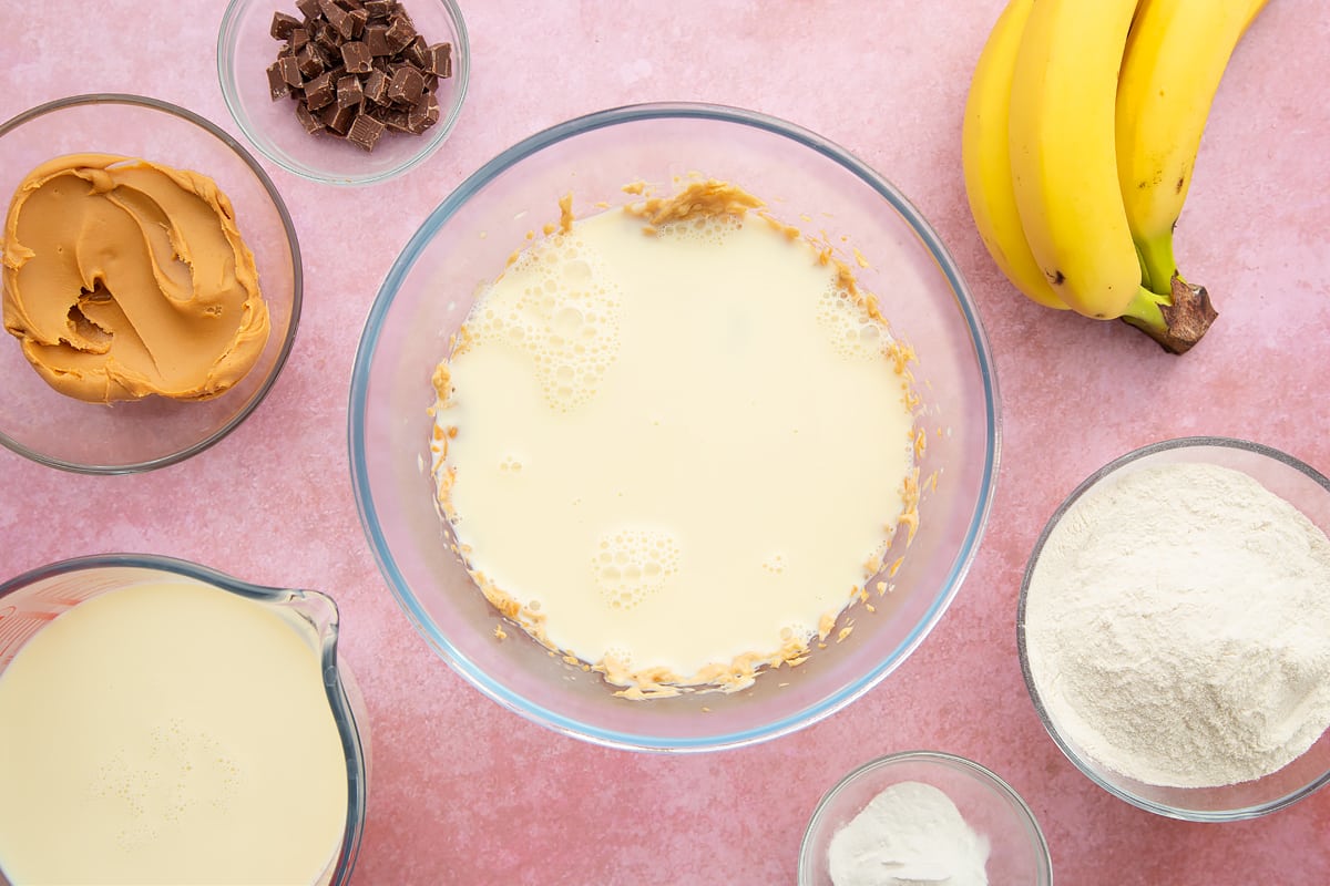 A bowl of banana and smooth peanut butter whisked together, topped with soya milk. The bowl is surrounded by the ingredients for vegan peanut butter pancakes.