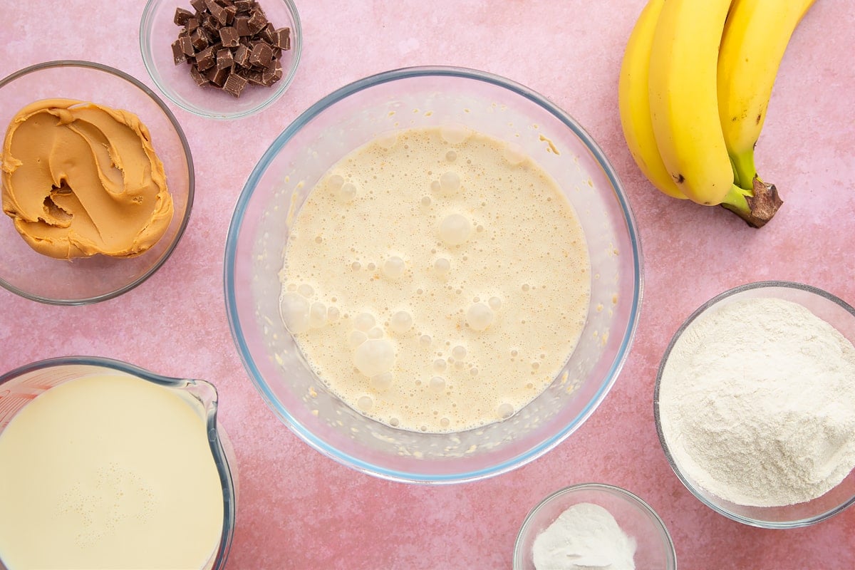 A bowl of banana, smooth peanut butter and soya milk whisked together. The bowl is surrounded by the ingredients for vegan peanut butter pancakes.