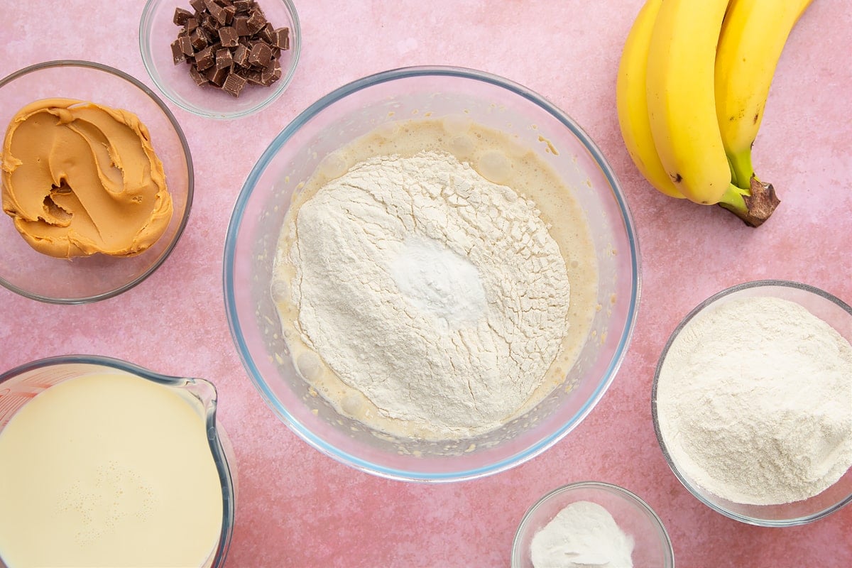 A bowl of banana, smooth peanut butter and soya milk whisked together, topped with flour and baking powder. The bowl is surrounded by the ingredients for vegan peanut butter pancakes.