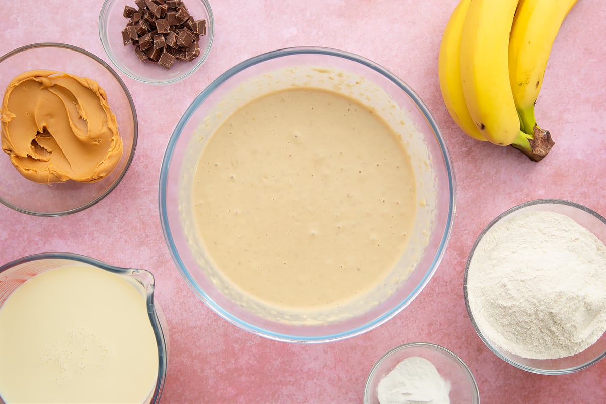 A bowl of banana, smooth peanut butter, soya milk, flour and baking powder whisked together. The bowl is surrounded by the ingredients for vegan peanut butter pancakes.