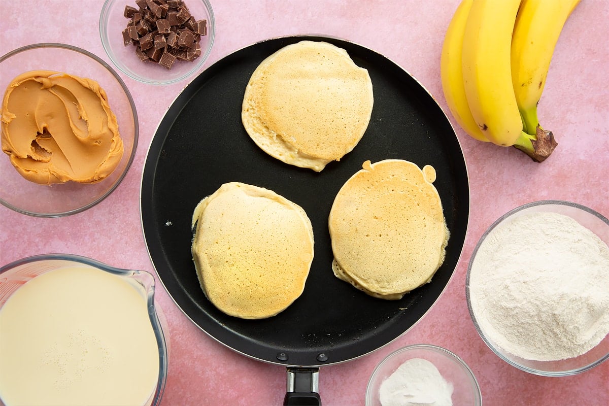 A frying pan with three vegan peanut butter and banana pancakes frying in it. The pan is surrounded by the ingredients for vegan peanut butter banana pancake batter.