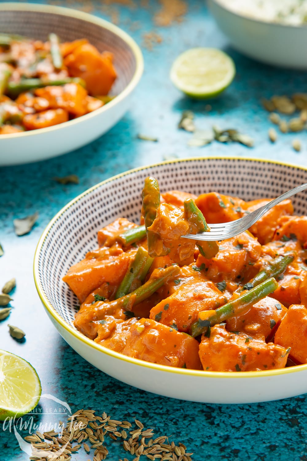 Vegan Sri Lankan curry in a yellow and black bowl. Another bowl is shown in the background and ingredients are scattered around it. A fork lifts some curry from the bowl.