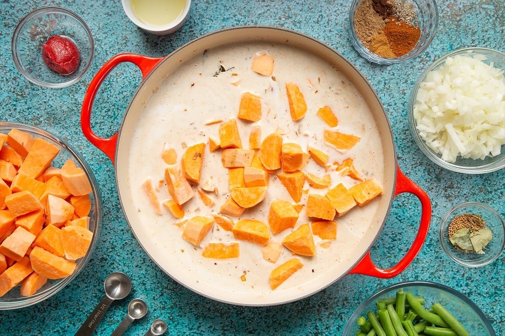 A large, shallow pan with sweet potato added to coconut milk, tomato paste, fried onion, ginger, garlic, chilli and spices. The pan is surrounded by ingredients for vegan Sri Lankan curry.