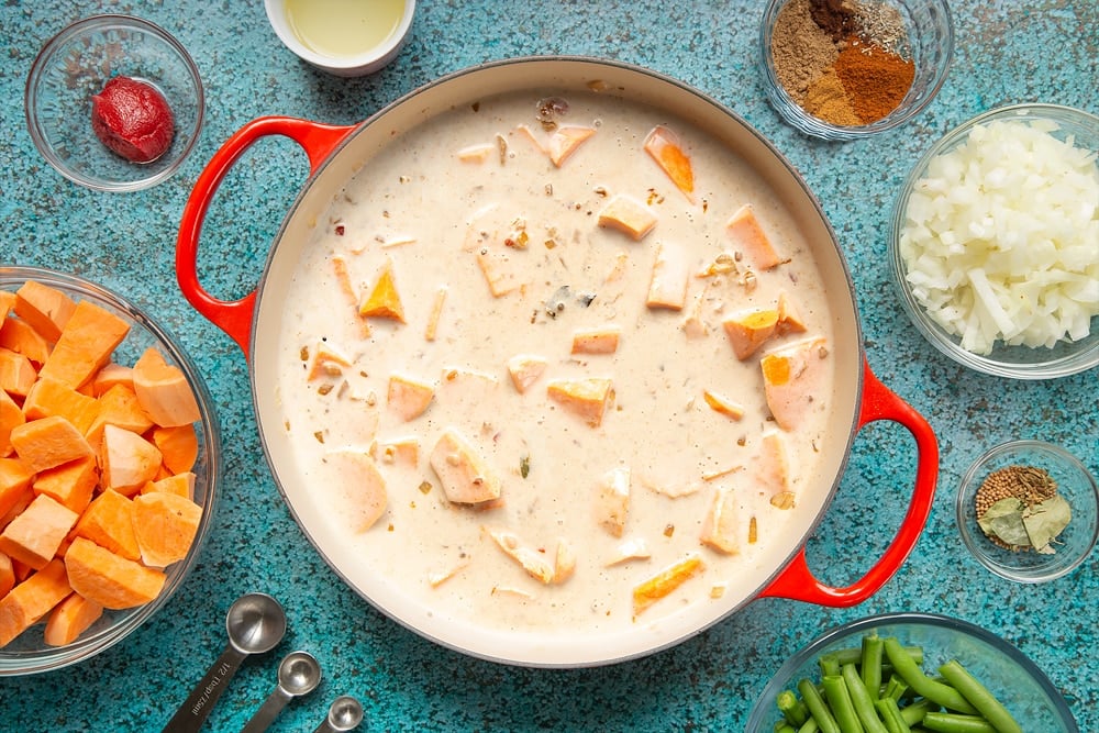 A large, shallow pan with sweet potato, coconut milk, tomato paste, fried onion, ginger, garlic, chilli and spices. The pan is surrounded by ingredients for vegan Sri Lankan curry.